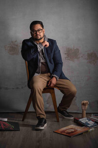 Man in a blue blazer and glasses seated on a wooden stool against a textured plaster wall, records at his feet.
