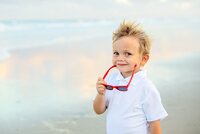Ponte Vedra portrait session capturing Young boy with wind-tousled blond hair holding red sunglasses and flashing a confident grin during a beach child portrait session.