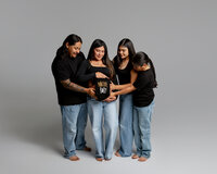 Family gathered around an expecting mom holding a “Native Baby” onesie over her belly during a maternity photoshoot in Yucaipa by Kaitlyn Dawn Photography.