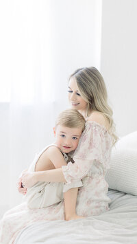 Light filled mommy and me portrait of a mom holding her toddler boy on her lap while he is snuggled with her. The photo was taken in a local photography studio in Dallas TX.