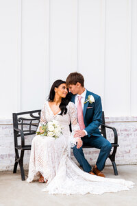 Newlyweds in their formal attire sitting on a bench outside of the white barn of Steel Magnolia Barn