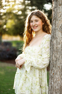 High school senior girl standing beside a tree in soft natural light wearing a floral dress, outdoor senior portrait by Kaitlyn Dawn Photography.