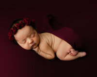 Sleeping newborn baby girl posed on a rich burgundy backdrop with a red floral headband during a studio newborn photography session by Yucaipa newborn photographer Kaitlyn Dawn Photography.
