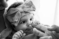 Professional photography by Nature's Child Photography: In Mandarin, a newborn with a large bow headband stares wide-eyed while clutching a parent’s finger, her tiny fingers wrapped tight during a lifestyle newborn.