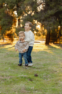 Older brother standing beside his toddler sibling in a sunlit park, candid sibling portrait photographed by Kaitlyn Dawn Photography, Yucaipa family photographer.