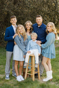 Outdoor family portrait of parents and four children standing together in coordinated blue outfits, natural light and greenery captured by Kaitlyn Dawn Photography, professional family photographer specializing in timeless outdoor portraits.