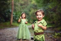 Jacksonville, FL portrait session capturing Smiling young boy in a green storybook-inspired outfit stands in the foreground as his sister in a flowing fairy dress holds a wand behind him during a fairy adventure signature session in Palatka.