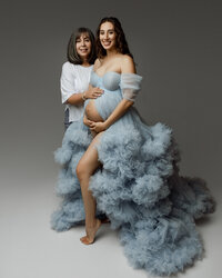 Pregnant woman smiling with her mother touching her belly, wearing a blue maternity gown during a heartfelt pregnancy session by Kaitlyn Dawn Photography in the Inland Empire.
