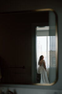 Bride in wedding gown looks out city window before wedding ceremony during quiet candid moment.