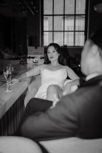 Mariée assise avec assurance au bar, robe bustier et coupe de champagne, marié de dos au premier plan, ambiance intime en noir et blanc chez Graziella