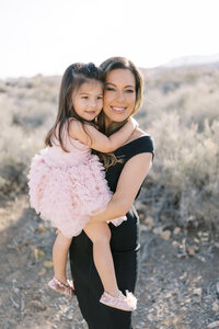 Mother from the Parker family holding her young daughter in a pink dress, both smiling in a warm desert setting in Las Vegas during a natural light family photography session.