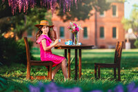 Young girl in bright pink dress seated at a small tea table beneath hanging purple wisteria