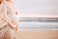 A close picture shows the belly of a pregnant mother peaking into frame with a shoreline and horizon in the background. It is an overcast day, but subtle pink hues can still be seen amongst the gray blue sky.