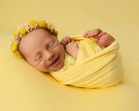 Smiling newborn baby wrapped in a bright yellow swaddle and wearing a delicate floral headband during a colorful newborn photography session by Kaitlyn Dawn Photography.