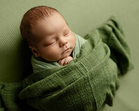 Sleeping newborn baby boy wrapped in a soft green swaddle and posed on a sage green backdrop during a studio newborn photography session by Kaitlyn Dawn Photography in Yucaipa.