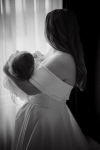 Mariée en robe bustier tenant son bébé blotti contre son épaule, lumière naturelle en contre-jour près d'une fenêtre au Château Vaudreuil, moment de tendresse en noir et blanc