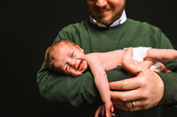 A smiling baby lies in her father's arms. He wears a green sweater and smiles down at her.