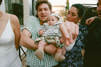 Parents holding their baby outdoors, smiling and pointing at camera