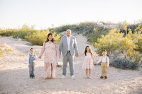 The Collins family standing together and holding hands in the Las Vegas desert, with parents and three children smiling in a bright, natural light outdoor family portrait session.