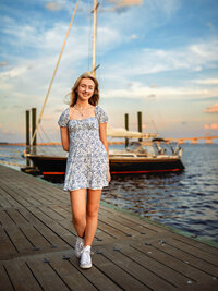 High school senior girl in floral dress walking on a dock with boat behind her in New Bern North Carolina