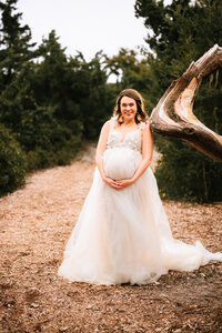 A pregnant woman leans against a curving tree branch. She stands on a mulched path with large bushes growing up behind her.