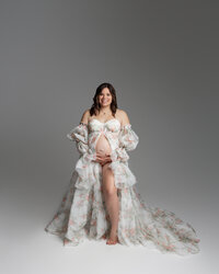 Pregnant woman smiling in a floral maternity gown during a soft studio maternity photography portrait by Kaitlyn Dawn Photography.