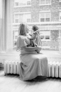 An indoor studio photo of a pregnant mom and toddler girl looking at snow outside