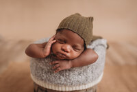Newborn baby boy, asleep,  propped on hands in basket with one hand on cheek and wearing a green sleepy cap