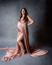 An expectant mother stands gracefully in a soft pink flowing maternity gown, her hand resting on her hip as she gazes confidently at the camera against a moody gray backdrop.