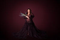 A pregnant woman in a dark plum gown holds a bouquet of dried lavender, her soft gaze and flowing gown creating a timeless, elegant look.