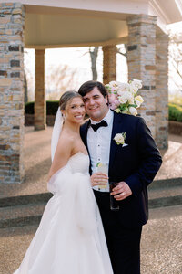 Bride and groom smiling and holding drinks while embracing in front of the stone pavilion at Burritt on the Mountain in Huntsville, Alabama, photographed by Perry Hancock.