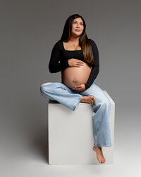 Pregnant mom sitting on a studio block in casual attire, softly holding her belly and looking off-camera during a maternity session in the Inland Empire by Kaitlyn Dawn Photography.