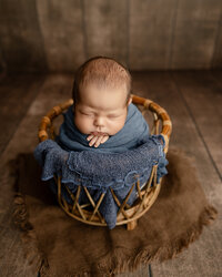 Sleeping newborn baby boy posed in a wicker basket with a blue wrap on a rustic wooden backdrop during a professional newborn photography session by Kaitlyn Dawn Photography in Yucaipa.