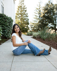High school senior girl sitting on a paved walkway with trees and greenery in the background, outdoor senior portrait by Kaitlyn Dawn Photography.