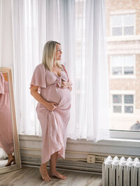 Pregnant woman standing by the window in front of the curtains, holding up her dress and holding her bump