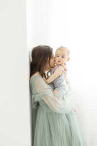 Photo of a mother leaning against a wall while she's holding her baby boy close to her in a DFW Photography studio.