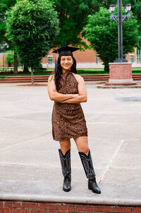 MTSU senior grad wearing black boots and standing on the bench outside campus library wearing leopard print dress