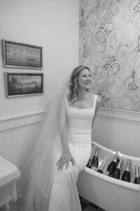 Bride in wedding dress sits on edge of bathtub filled with champagne bottles.