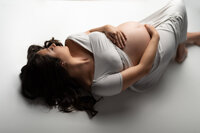 Pregnant woman in a soft gray gown lying on a seamless white studio floor, gently holding her belly with eyes closed during a peaceful, minimalist maternity session in Waukesha near Hartland, Wisconsin.