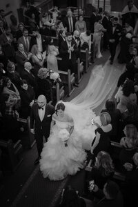 Mariée marchant dans l’allée d’une église aux côtés d’un homme en smoking, entourés de proches émus, sous une lumière douce et poétique, capturant l’intensité et la tendresse du moment en noir et blanc.