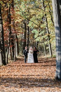 A bride, wearing a long veil and holding a bouquet, walks down a leaf-covered forest aisle