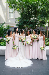 A bridal party wearing soft pink gowns smiles and laughs with their friend, the bride, while posing for bridal party portraits at their DMV wedding