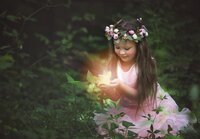 In O’Fallon, Illinois, a young girl in a flower crown smiles softly as she holds a glowing fairy in her hands during a signature fairy adventure. - A portrait experience by Nature's Child Photography.