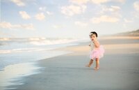 Baby girl in a pink tutu standing barefoot at the water’s edge, looking surprised as waves roll in during a beach child photography session- A portrait experience by Nature's Child Photography.