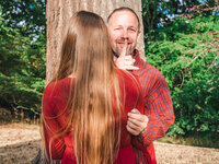 Man secretly holds an engagement ring behind his back and makes a shh gesture while standing with his girlfriend during a proposal photo session in a Los Angeles park.