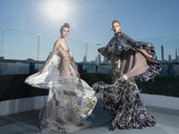 Two models in elaborate printed gowns on a rooftop terrace, sun flaring behind them above a city skyline.