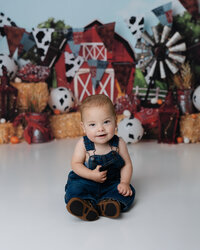 Smiling baby boy sitting on the studio floor in front of a barnyard-themed cake smash backdrop with hay bales and cow print balloons.
Photograph by Yucaipa cake smash photographer Kaitlyn Dawn Photography.