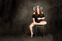 A pregnant woman in a black bodysuit sits on a black stool. She is on a gray painted backdrop. She has one leg propped up on the stool and the other draped to the side with her toes touching the ground. She wears a black and white striped hat and has long brown hair.