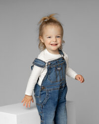 Smiling toddler girl standing on white studio steps against a soft gray backdrop, wearing denim overalls and a white top, classic studio child portrait captured by Kaitlyn Dawn Photography, professional family and children’s photography studio.