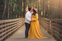 Pregnant mother in mustard yellow gown standing on wooden bridge surrounded by trees during maternity session